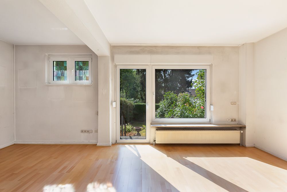 Helles Zimmer mit Parkettboden, großen Fenstern und Blick auf einen Garten.