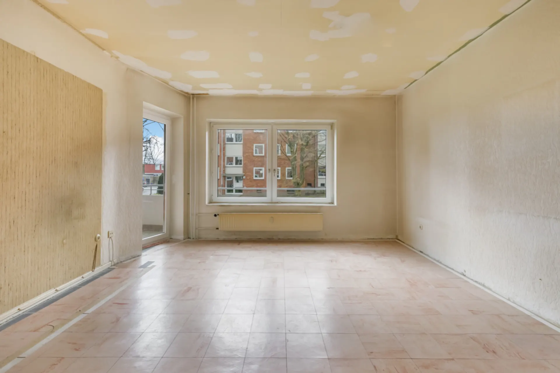 Leerer Raum mit großem Fenster, Tür zum Balkon und sichtbarer Heizung unter dem Fenster.
