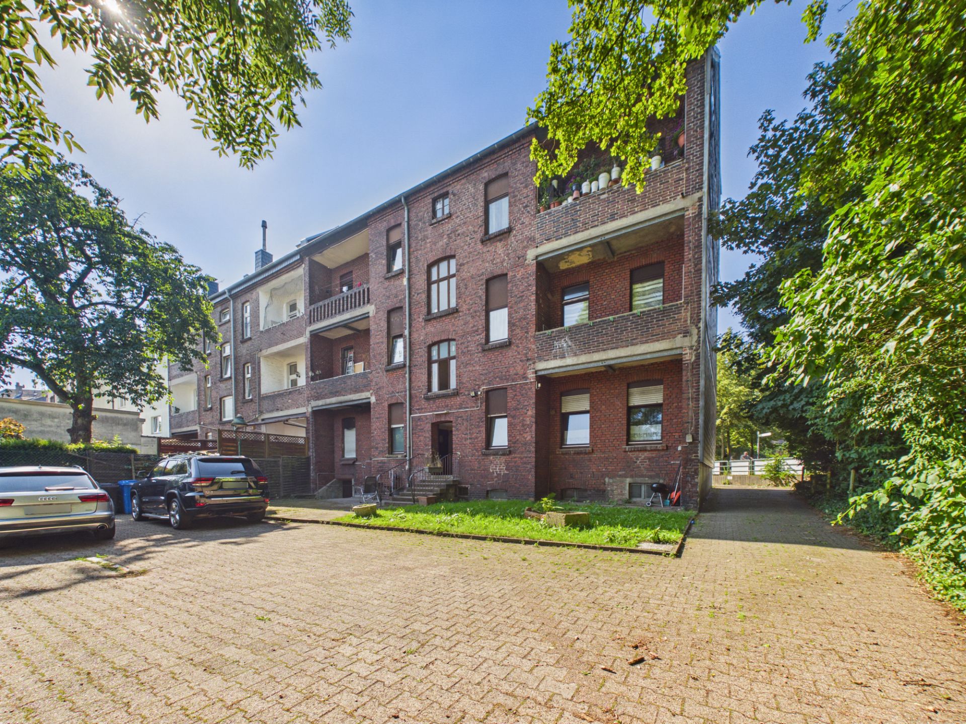 Backsteinwohnhaus mit Balkonen und kleinem Parkplatz im Vordergrund, umgeben von Bäumen.