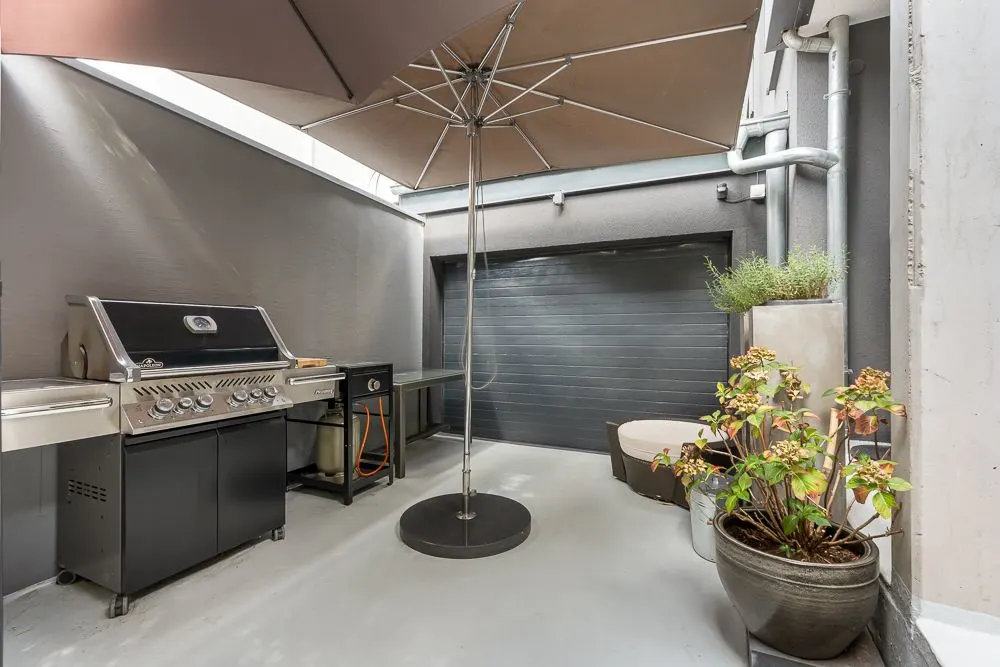 Kleine Terrasse mit Grill, großem Sonnenschirm, Blumentöpfen und grauer Wand.
