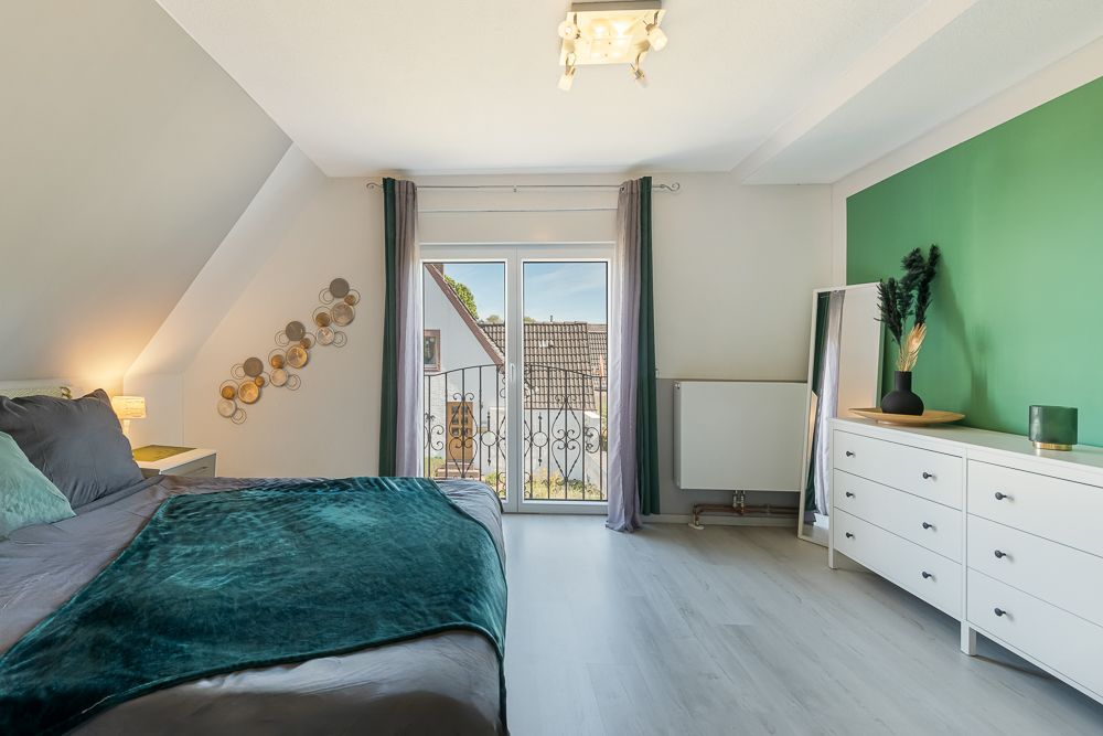Helles Schlafzimmer mit Doppelbett, Kommode, Spiegel und Balkonfenster.