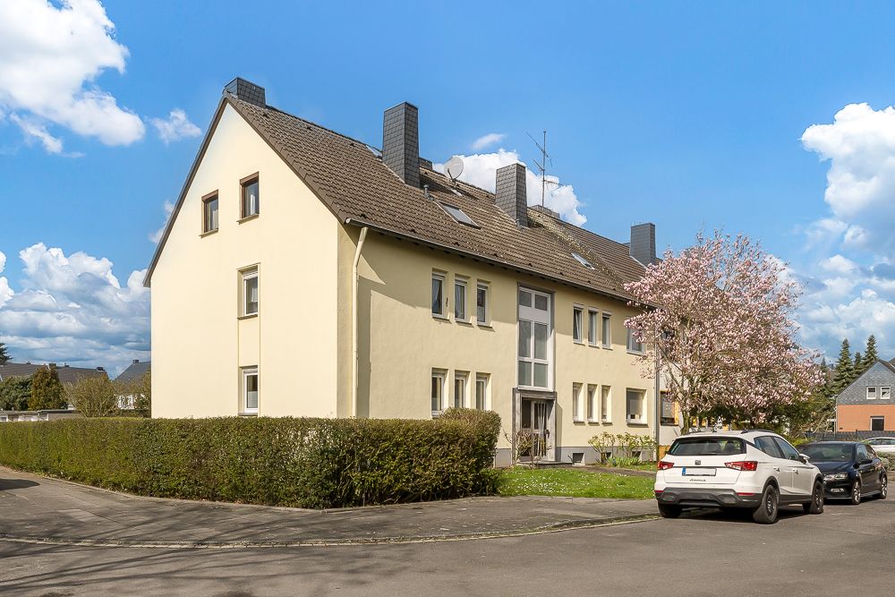 Dreistöckiges Wohnhaus mit hellem Putz, blühendem Baum und geparkten Autos am Straßenrand.