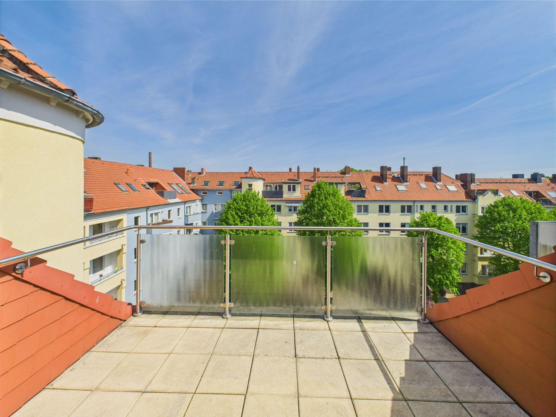 Balkon mit Glasgeländer und Blick auf Wohnhäuser mit roten Dächern und Bäumen.