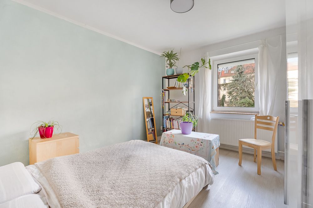 Helles Schlafzimmer mit Bett, Regal, Stuhl und Pflanzen am Fenster.