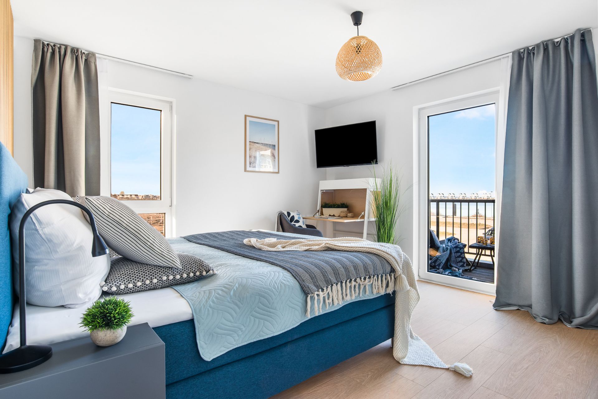 Modernes Schlafzimmer mit blauem Bett, zwei Fenstern, Balkon und Fernseher an der Wand.