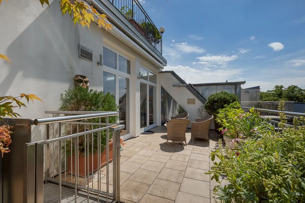 Große Dachterrasse mit Pflanzen, zwei Stühlen und Zugang zu einem modernen Wohngebäude bei Sonnenschein.