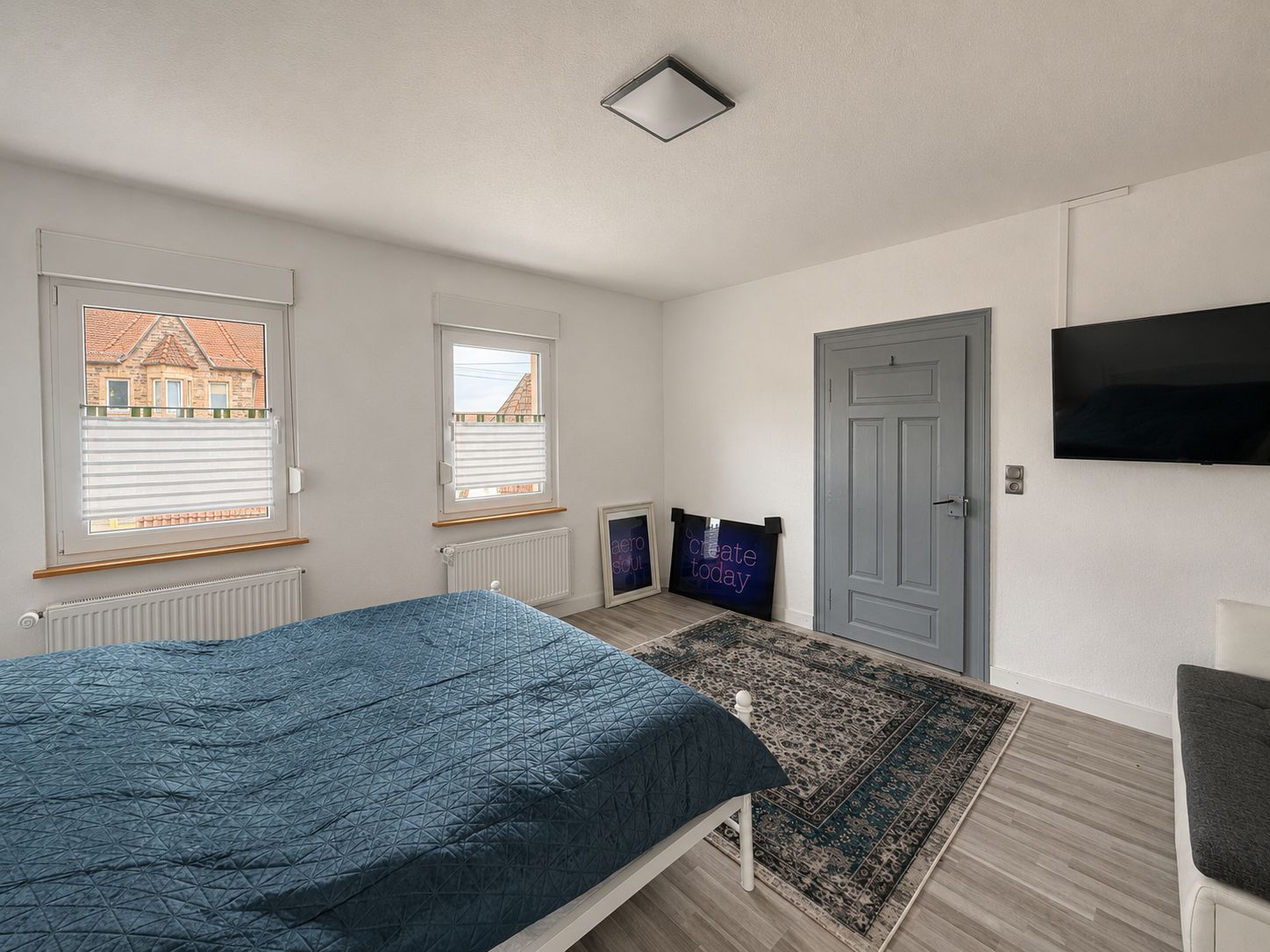 Helles Schlafzimmer mit Bett, Teppich, zwei Fenstern, grauer Tür und Wandfernseher.