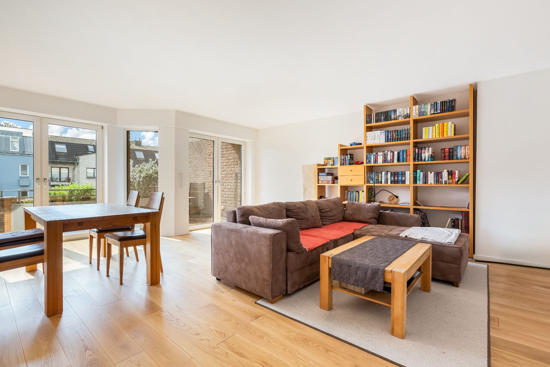 Helles Wohnzimmer mit Ecksofa, Bücherregal, Esstisch und großen Fenstern.