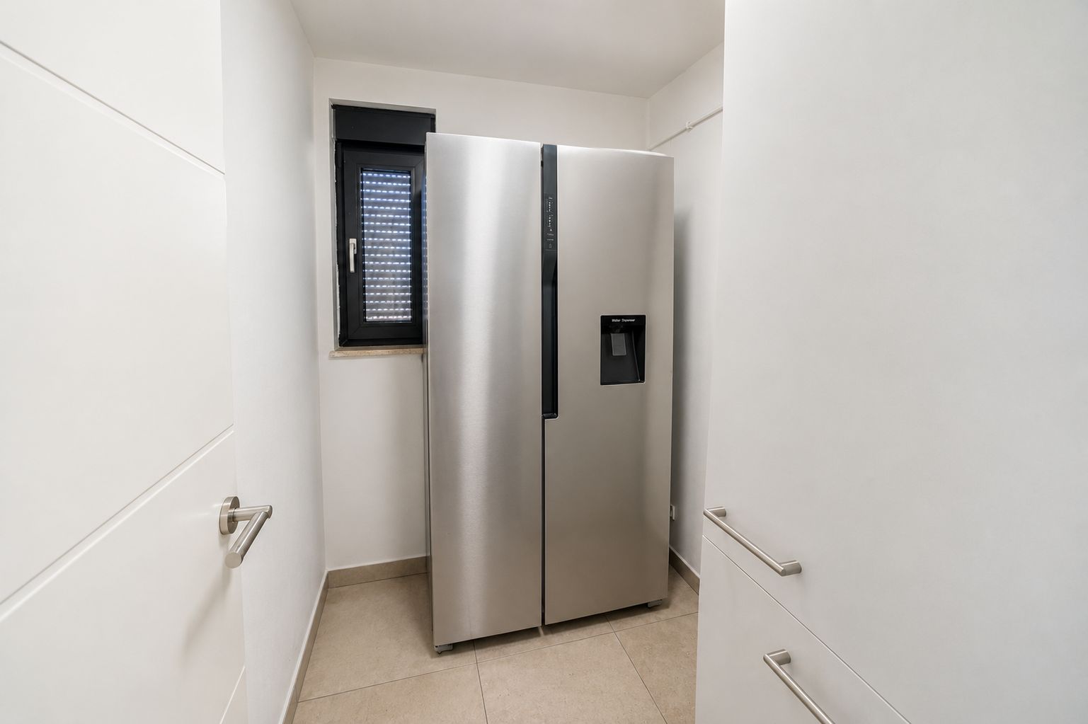 Ein moderner silberner Kühlschrank steht in einem kleinen, weißen Raum mit Fenster.