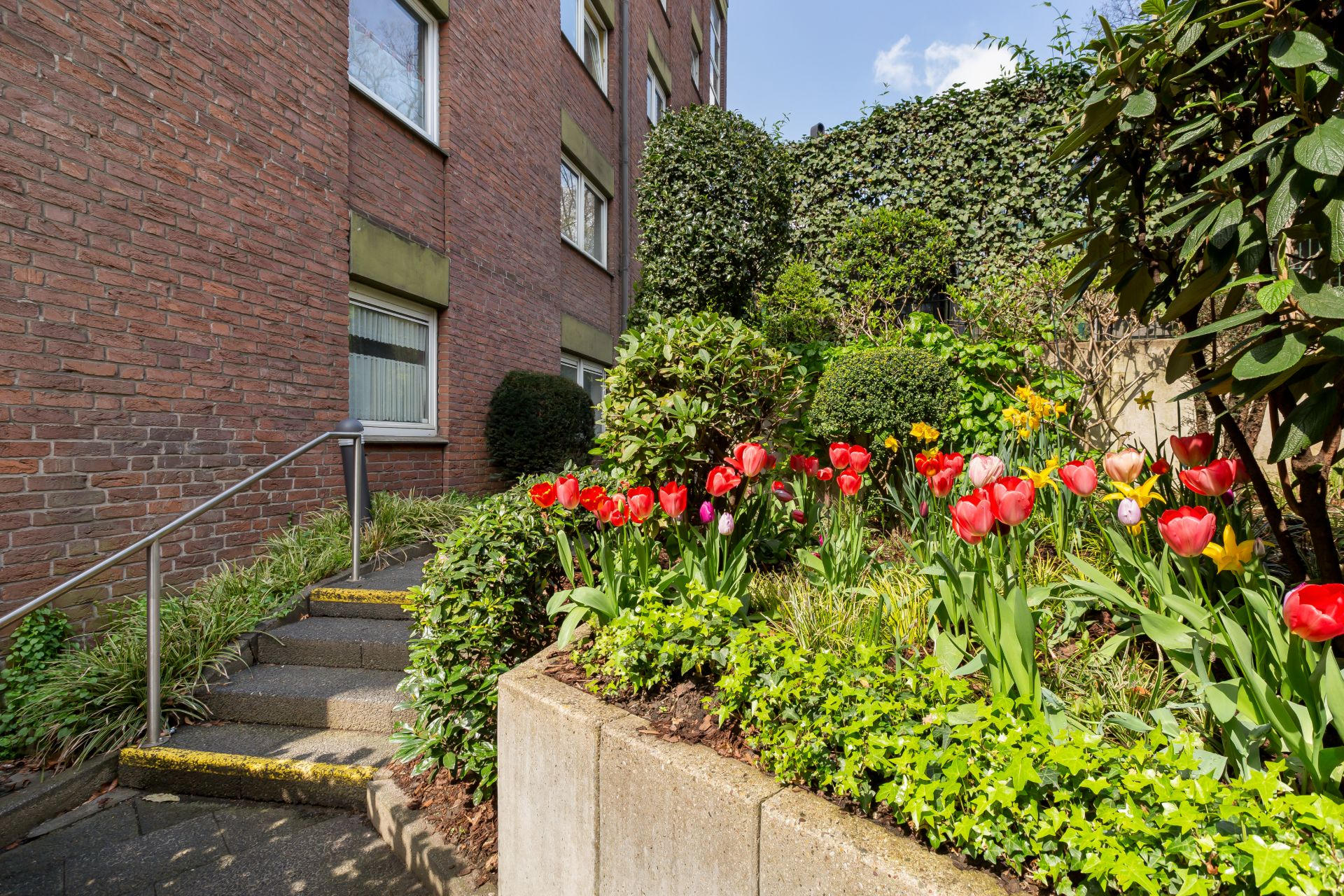 Treppe neben einem Blumenbeet mit Tulpen vor einem Wohngebäude aus Backstein.
