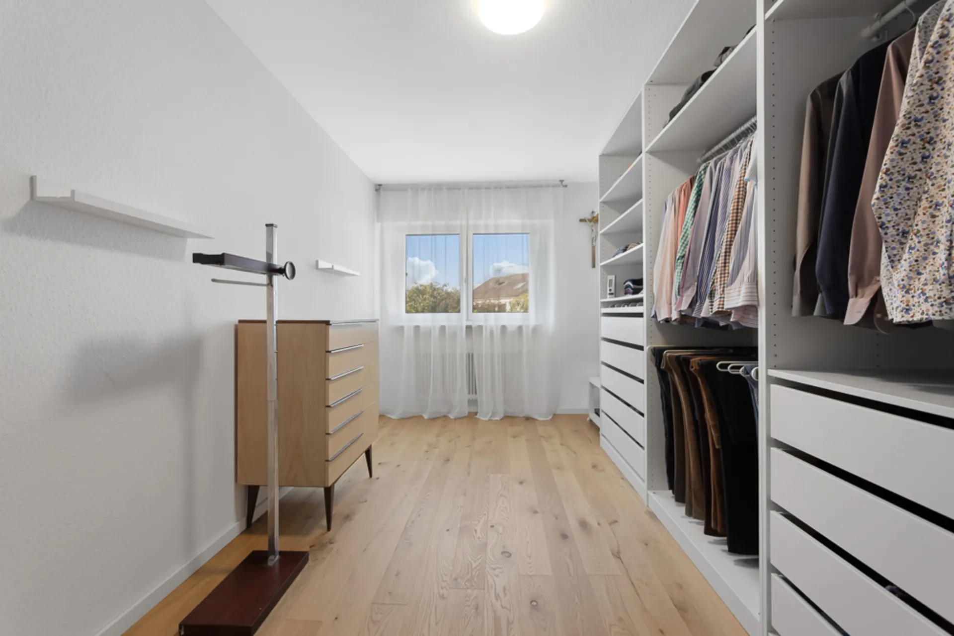 Heller begehbarer Kleiderschrank mit Holzschrank, Regalen und Kleidung, Fenster mit Vorhang am Ende.
