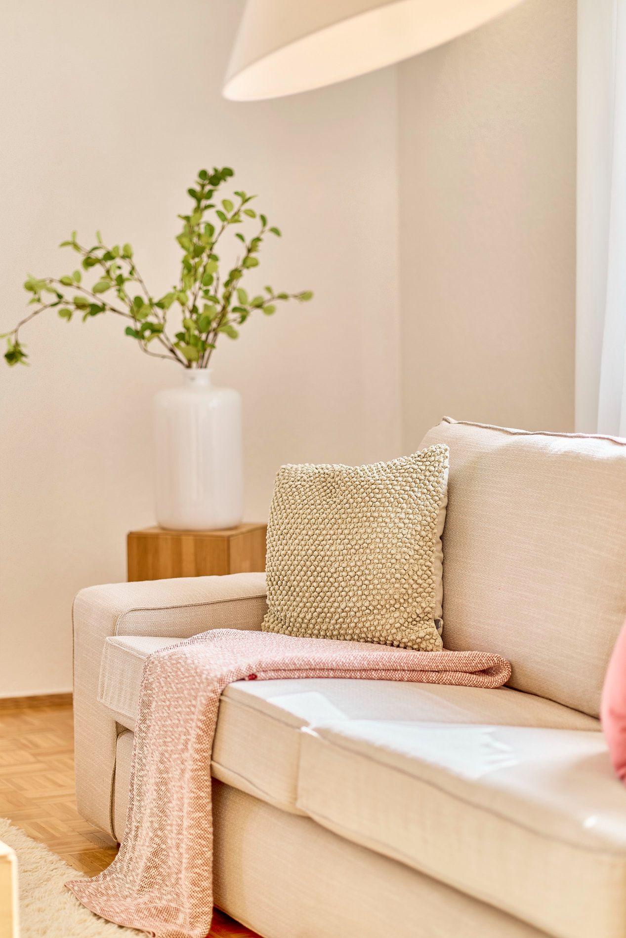 Beiges Sofa mit Kissen und rosa Decke, daneben ein Tisch mit Vase und grünen Zweigen.
