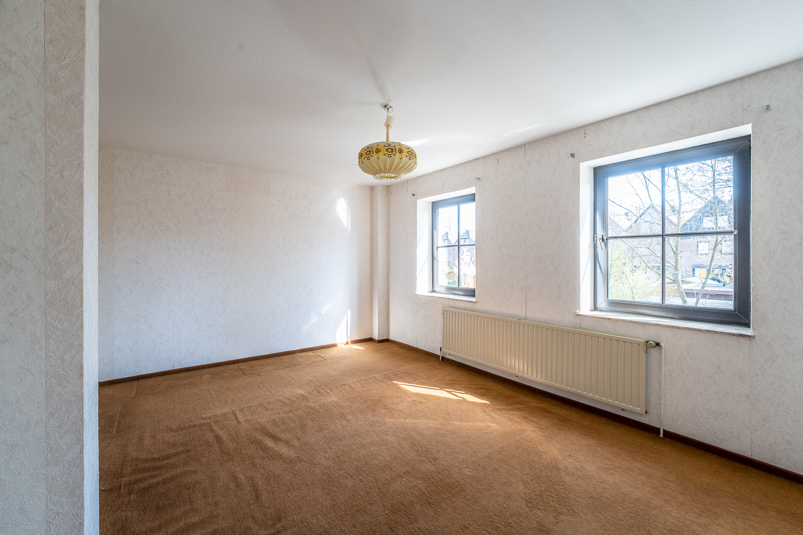 Leeres Zimmer mit braunem Teppich, zwei Fenstern und heller Deckenlampe.