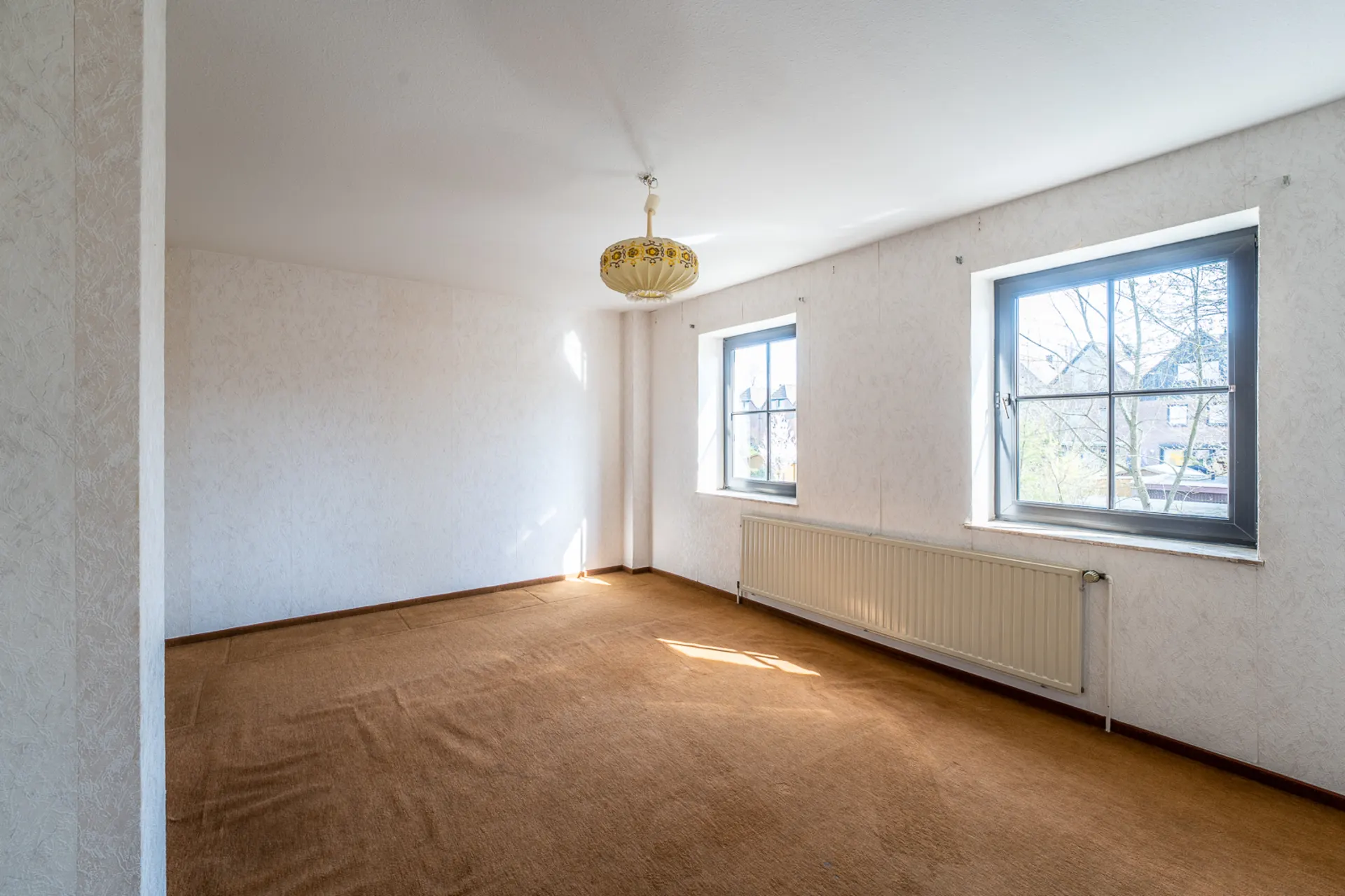 Leeres Zimmer mit braunem Teppich, zwei Fenstern und heller Deckenlampe.