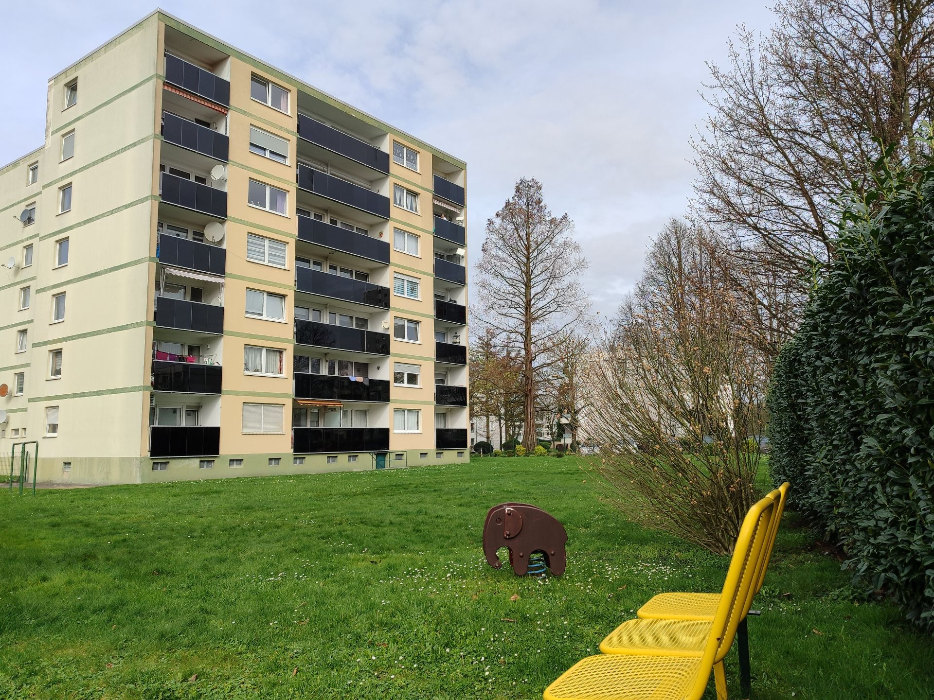 Mehrfamilienhaus mit Balkonen, davor Wiese, Spieltier und zwei gelbe Bänke.