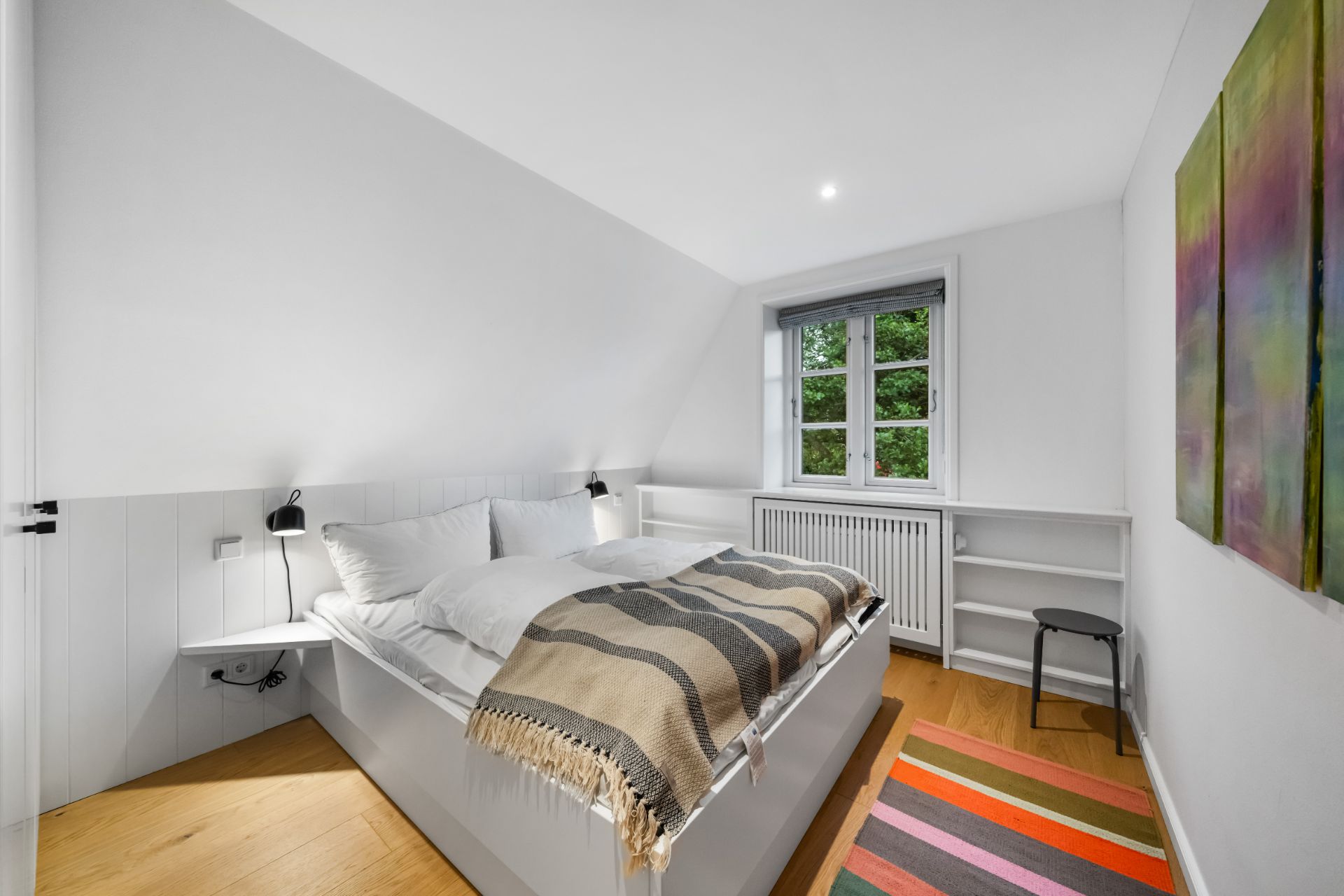 Helles Schlafzimmer mit Doppelbett, farbigem Teppich, Fenster und Wandbild.