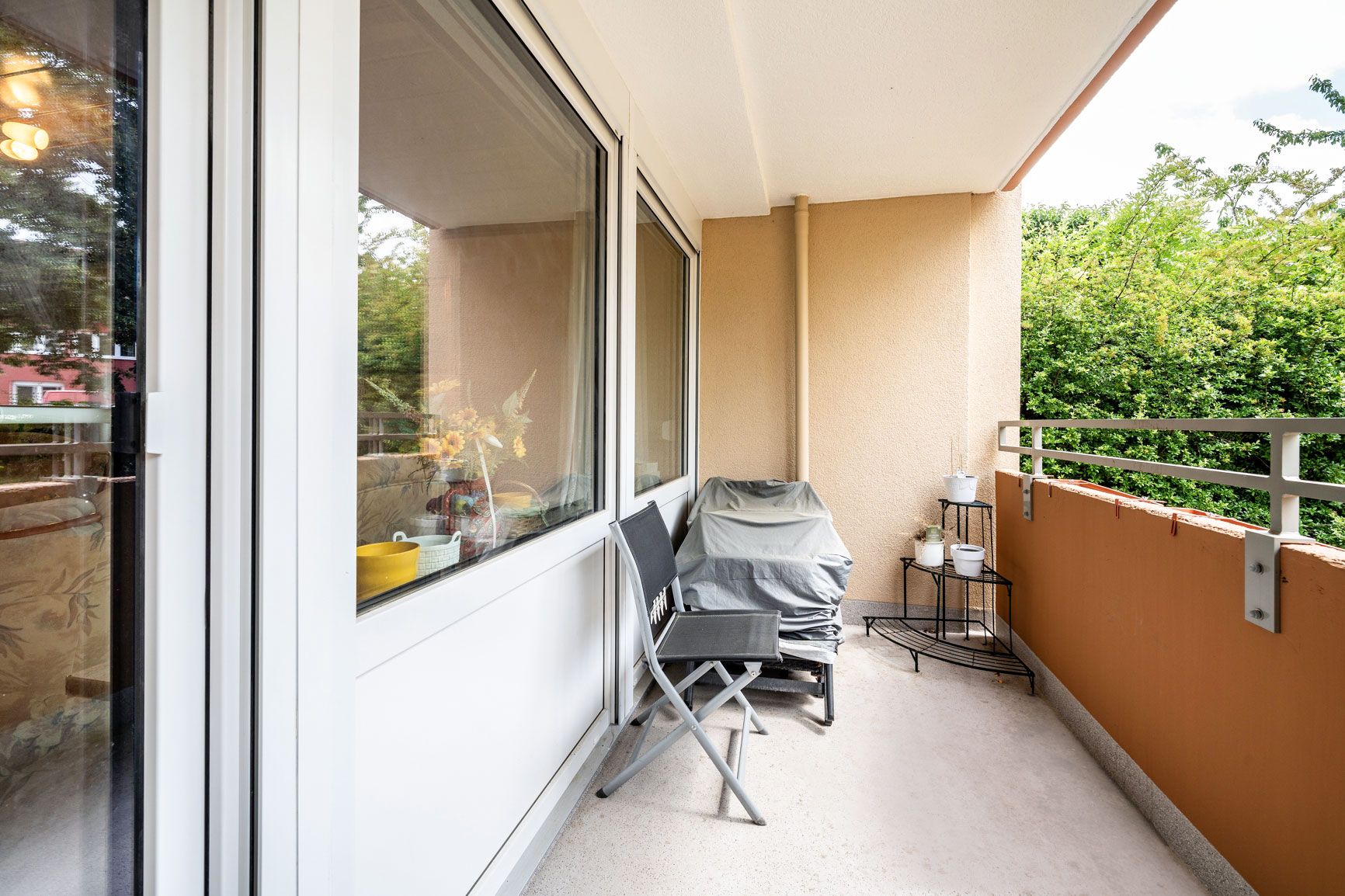 Balkon mit zwei Stühlen, einem abgedeckten Grill und Pflanzenregal, umgeben von grünem Bewuchs.