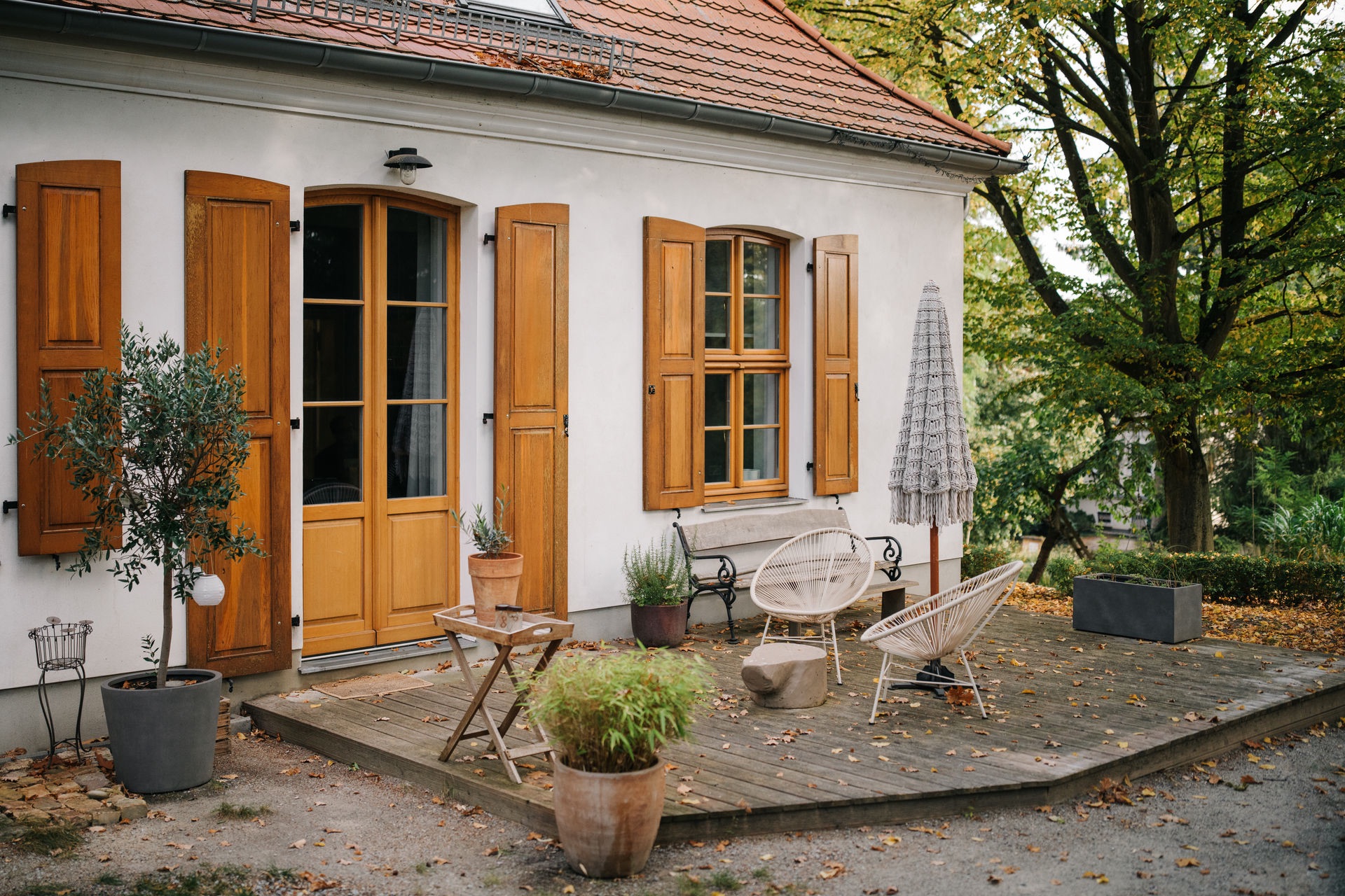 Holzterrasse mit Pflanzen, Stühlen, Tisch und Sonnenschirm vor einem Haus mit braunen Fensterläden.