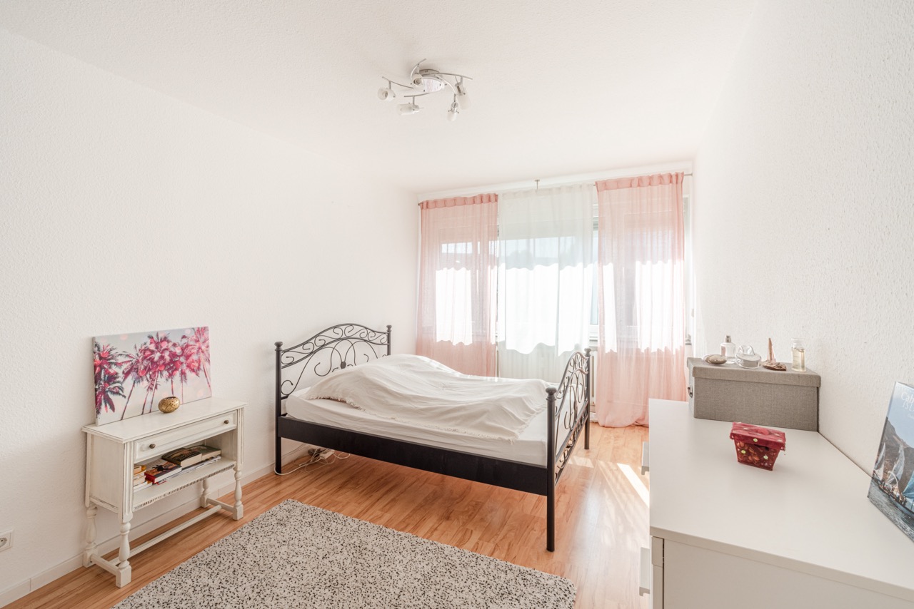 Helles Schlafzimmer mit Doppelbett, weißer Kommode, Teppich und rosa Vorhängen am Fenster.