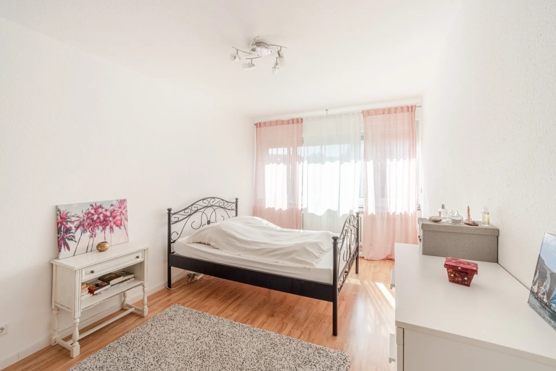 Helles Schlafzimmer mit Doppelbett, weißer Kommode, Teppich und rosa Vorhängen am Fenster.