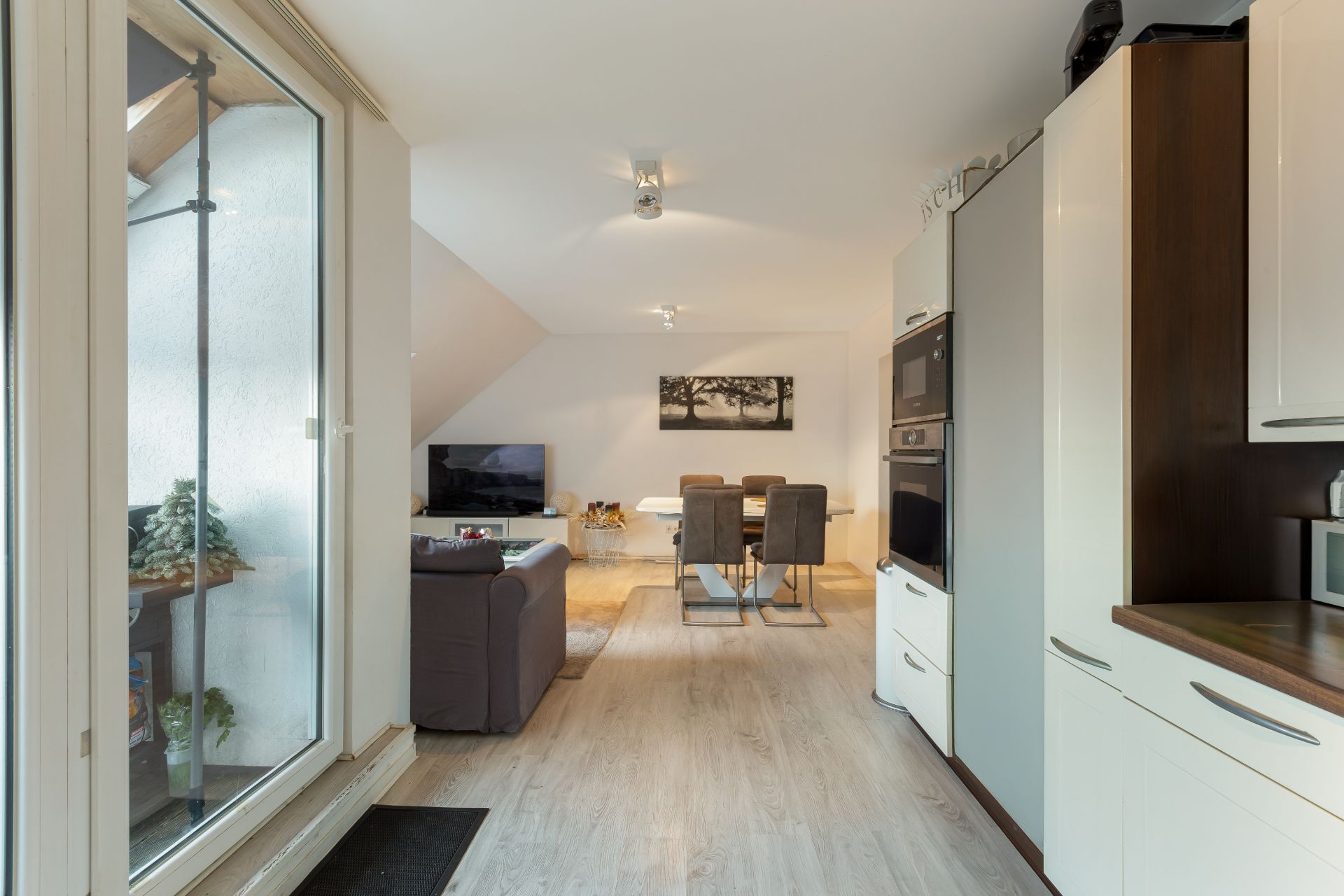 Helles Wohnzimmer mit Sofa, Esstisch und offener Küche, große Fenster links.