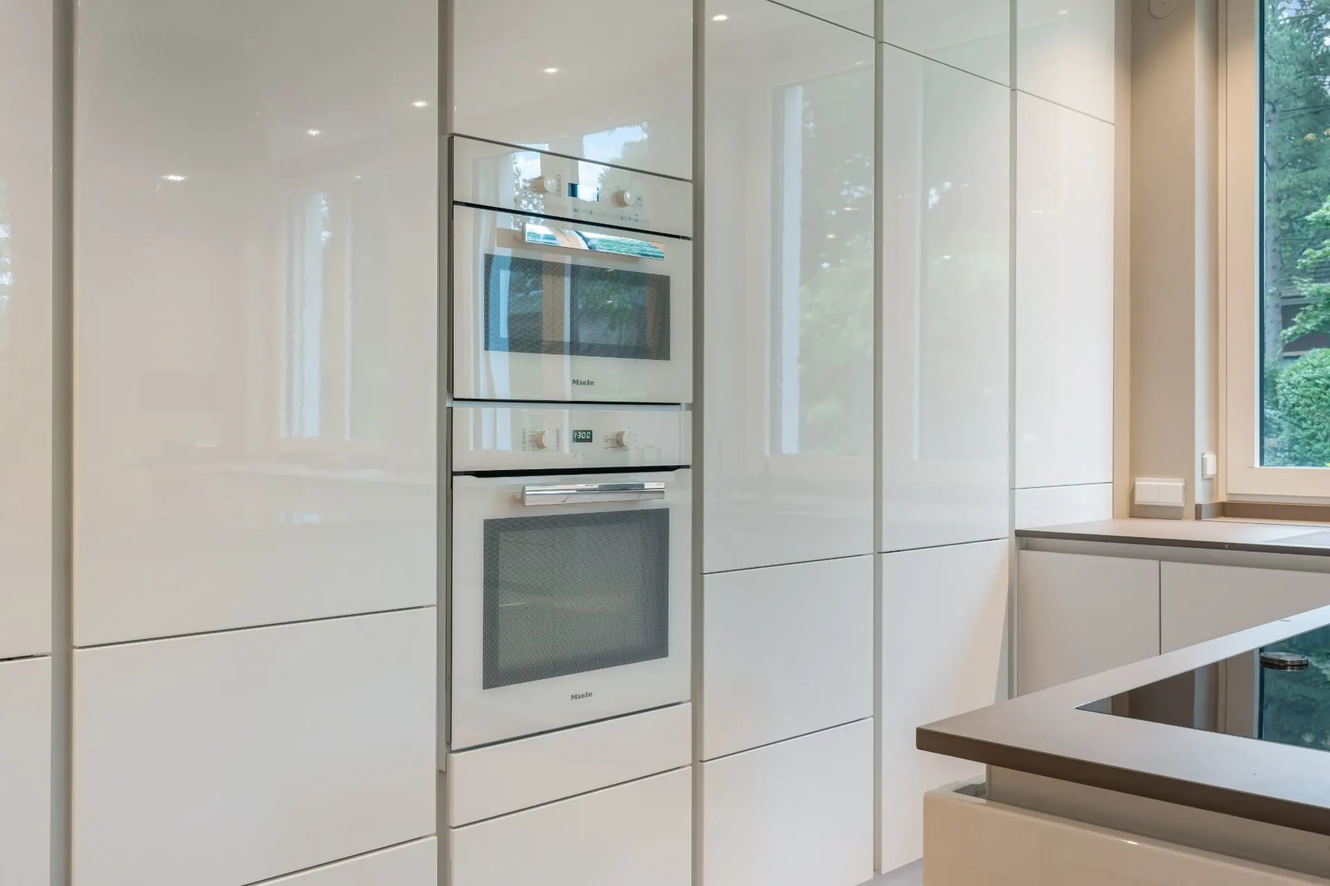 Moderne Küche mit weißen Hochglanzschränken und Einbaugeräten neben einem Fenster.