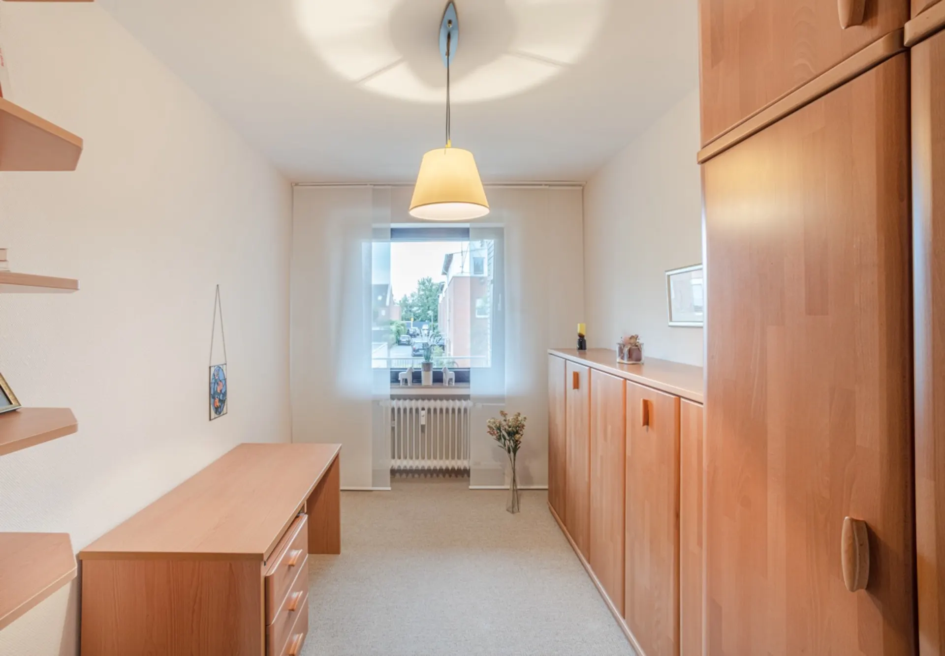 Helles Zimmer mit Holzschreibtisch, Regalen, Schränken und Fenster mit Vorhang.