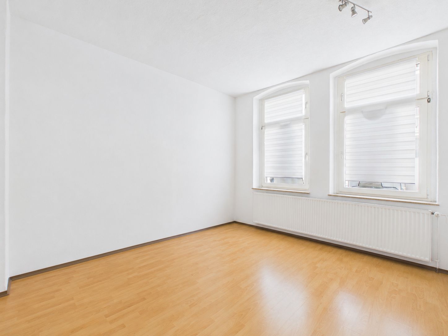 Heller, leerer Raum mit zwei Fenstern, weißer Wand und Laminatboden.