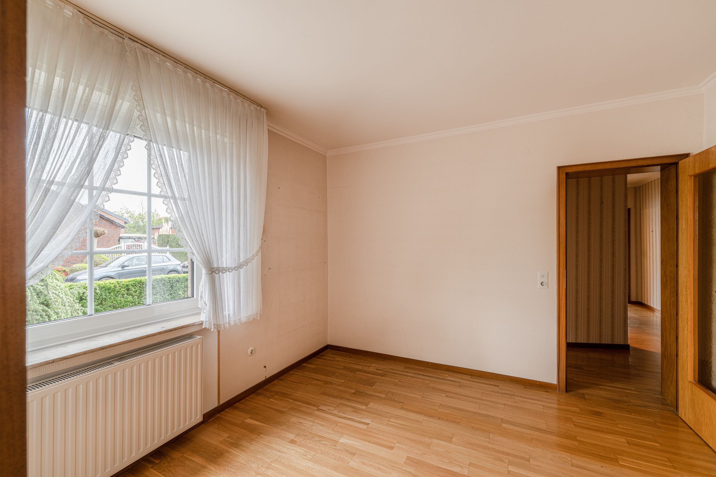 Helles, leeres Zimmer mit Holzfußboden, großem Fenster und weißen Gardinen.