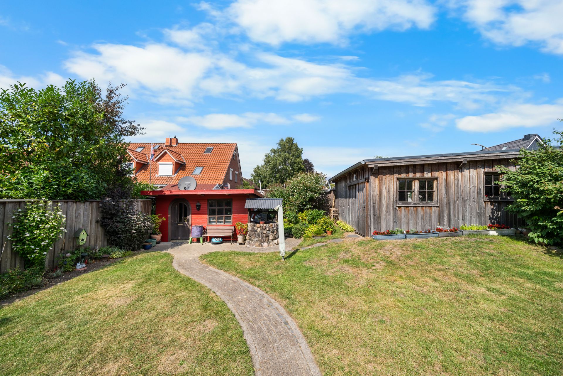 Garten mit Rasen, Weg, rotem Haus, Holzschuppen und Bäumen bei blauem Himmel.
