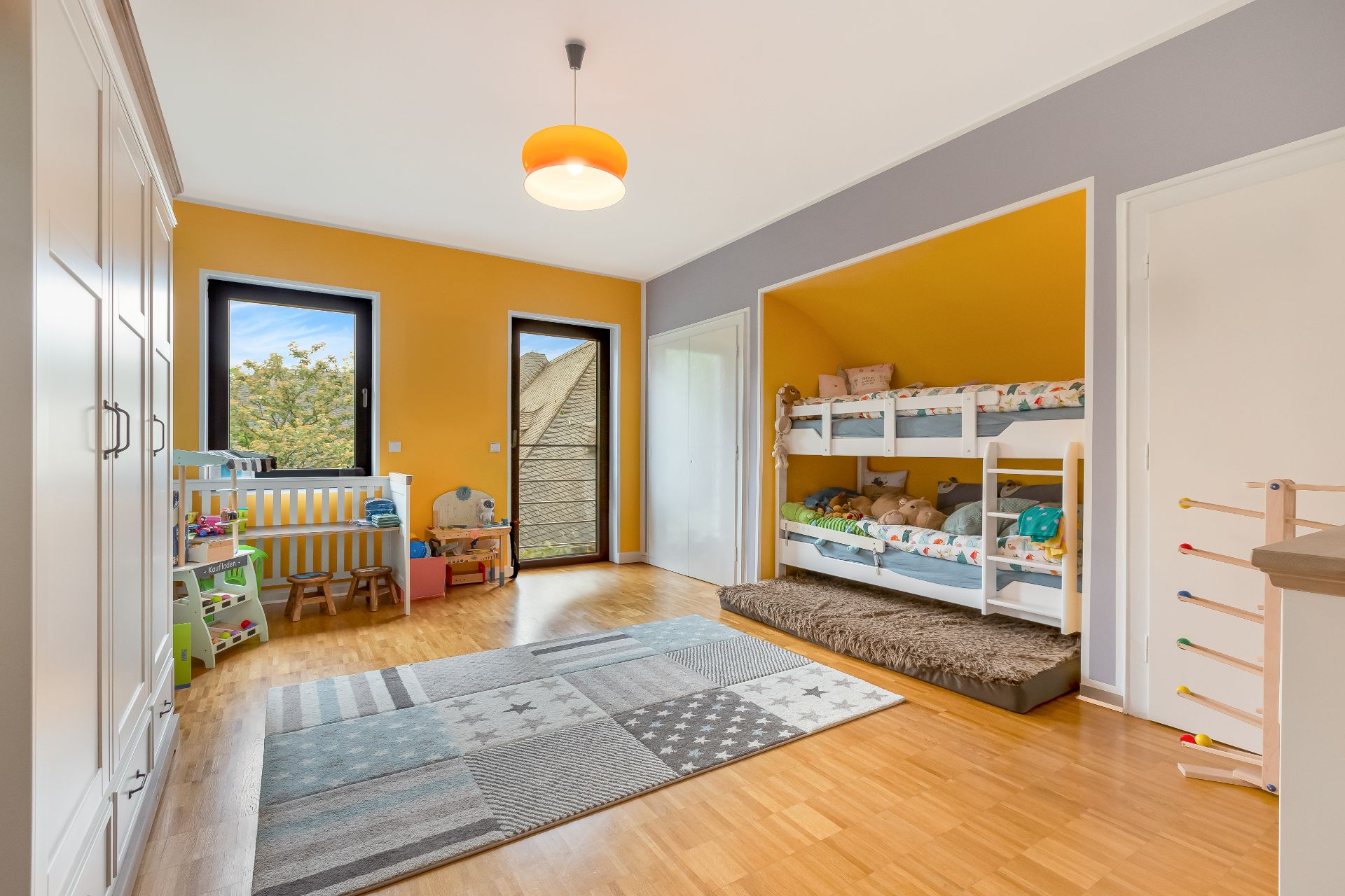 Helles Kinderzimmer mit Etagenbett, Teppich, Spielzeug und Kinderbett vor gelber Wand.