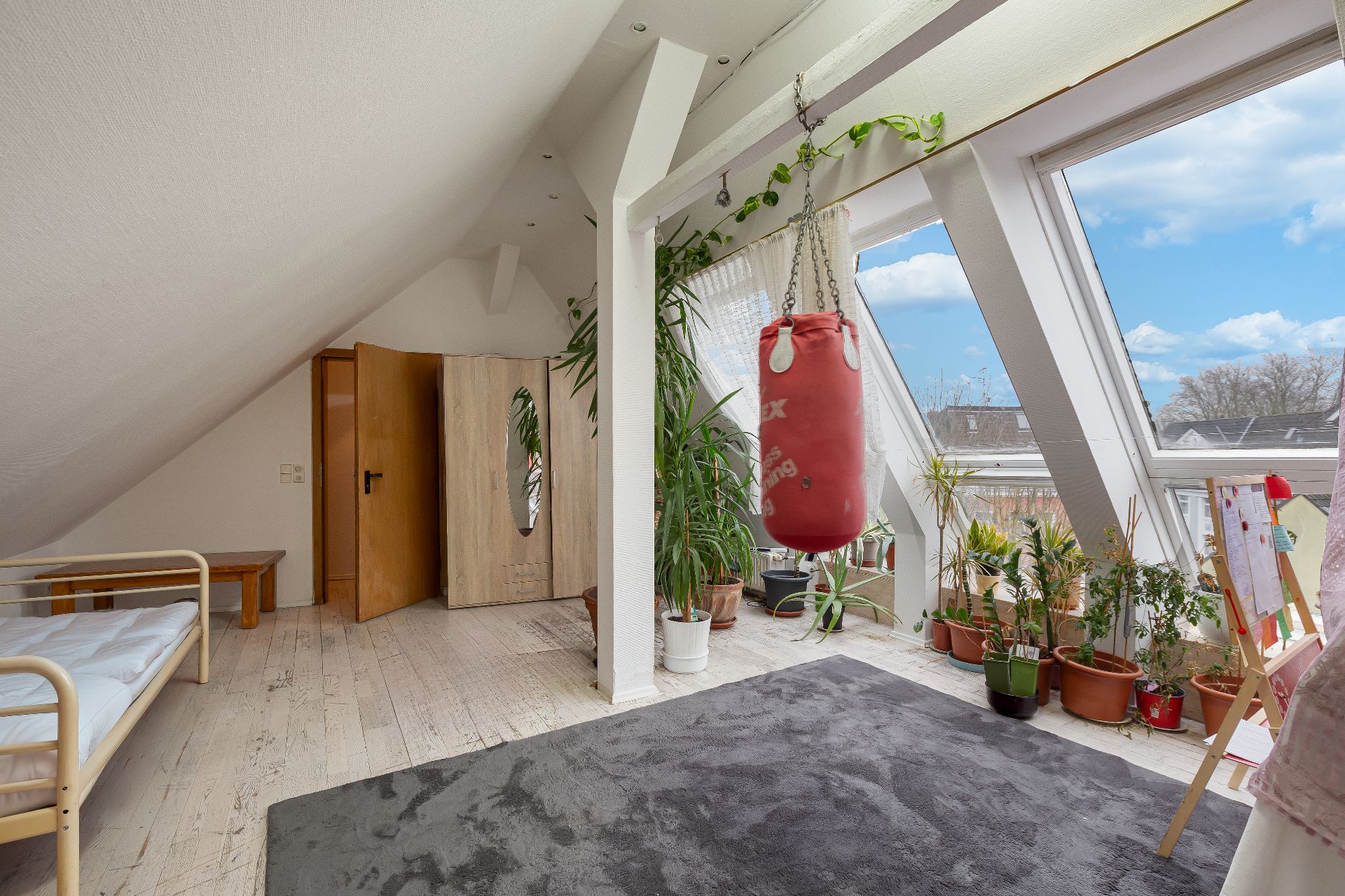 Helles Zimmer mit Bett, Hängeboxsack, vielen Pflanzen und großen Fenstern.