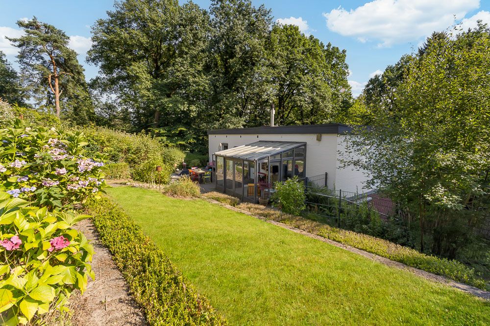Haus mit Wintergarten und Garten, umgeben von Bäumen und blühenden Sträuchern.