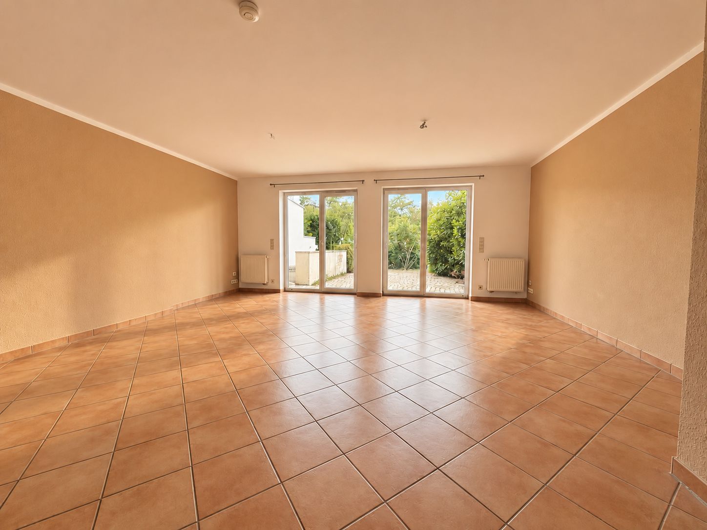 Leerer Raum mit Fliesenboden und großen Fenstern, die auf eine Terrasse mit Gartenblick führen.
