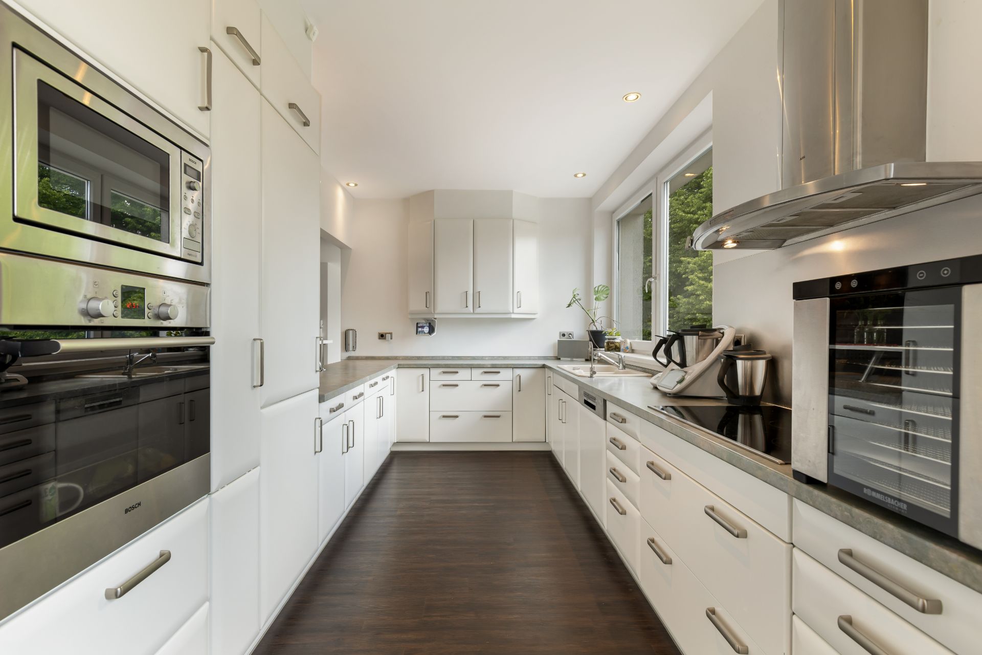 Moderne Küche mit weißen Schränken, Einbaugeräten und dunklem Boden, große Fenster rechts.