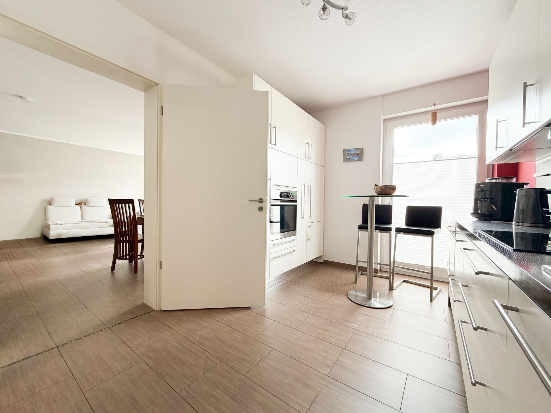 Helle moderne Küche mit weißen Schränken, kleinem Esstisch und Blick ins Wohnzimmer mit Sofa.