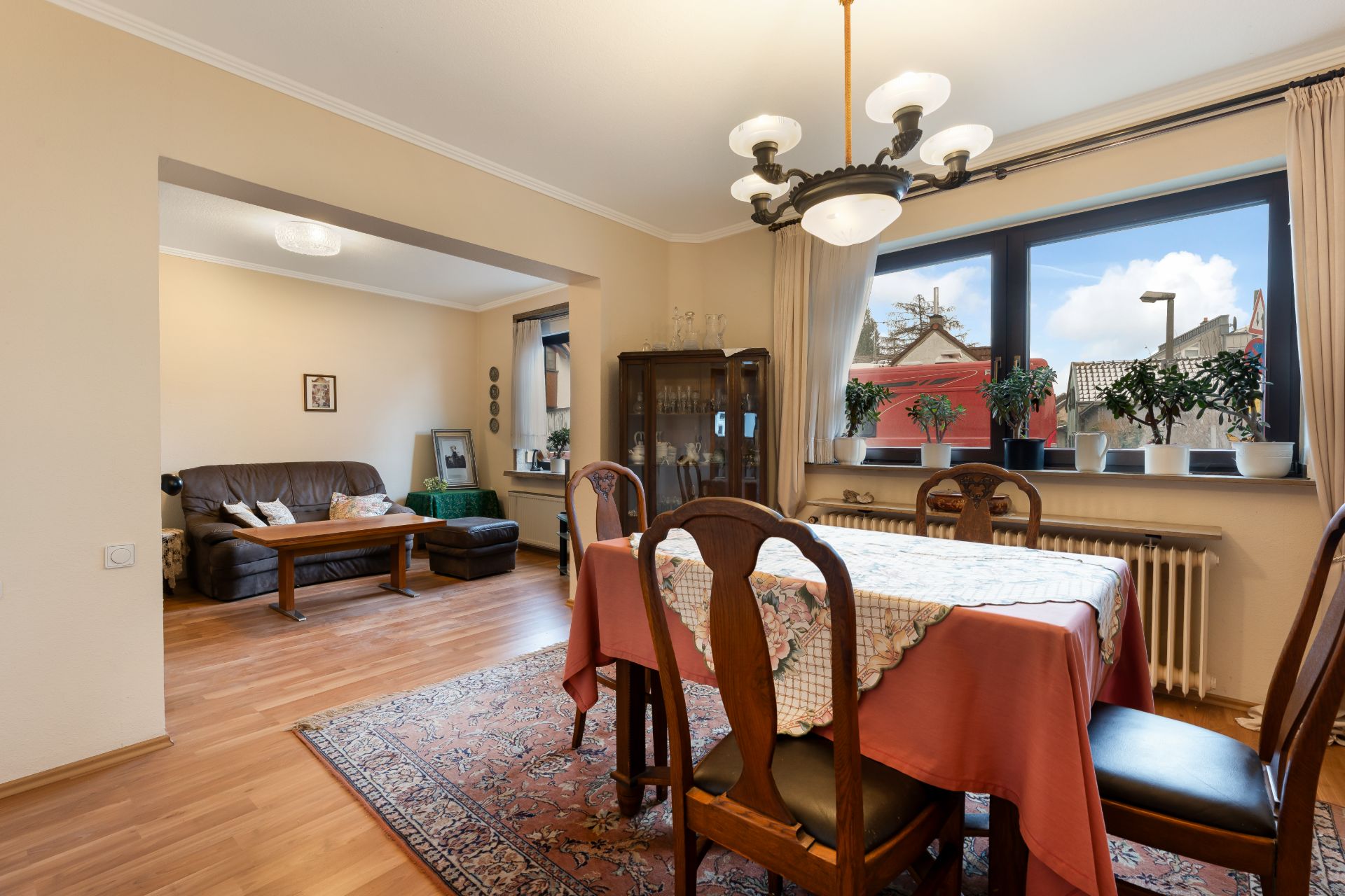Esszimmer mit Holztisch, sechs Stühlen, Teppich und Blick ins Wohnzimmer, Fenster mit Pflanzen.