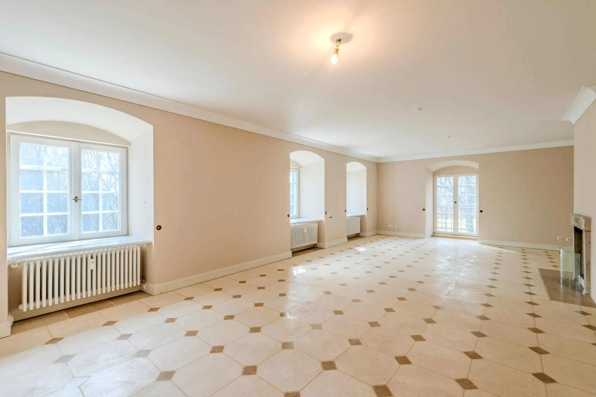 Heller, leerer Raum mit Fliesenboden, drei Fenstern, Glastür und Kamin.