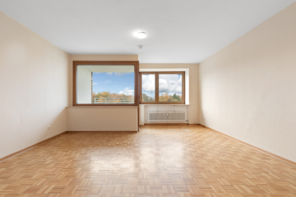 Leeres Wohnzimmer mit Parkettboden, großen Fenstern und Heizkörper unter dem Fenster.