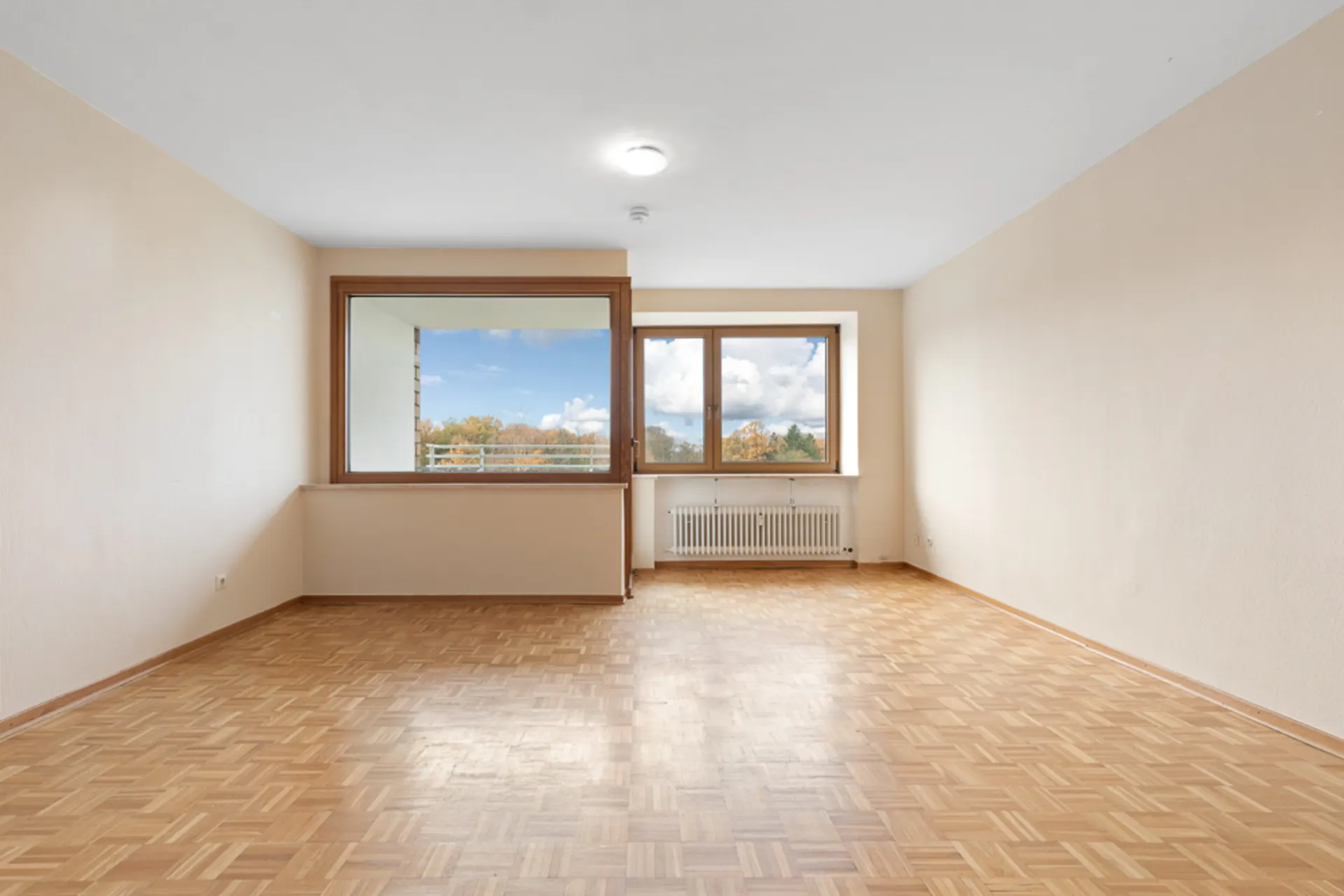 Leeres Wohnzimmer mit Parkettboden, großen Fenstern und Heizkörper unter dem Fenster.