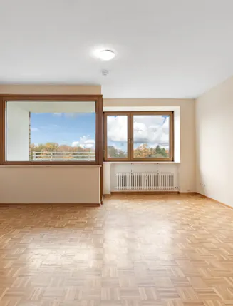 Leeres Wohnzimmer mit Parkettboden, großen Fenstern und Heizkörper unter dem Fenster.