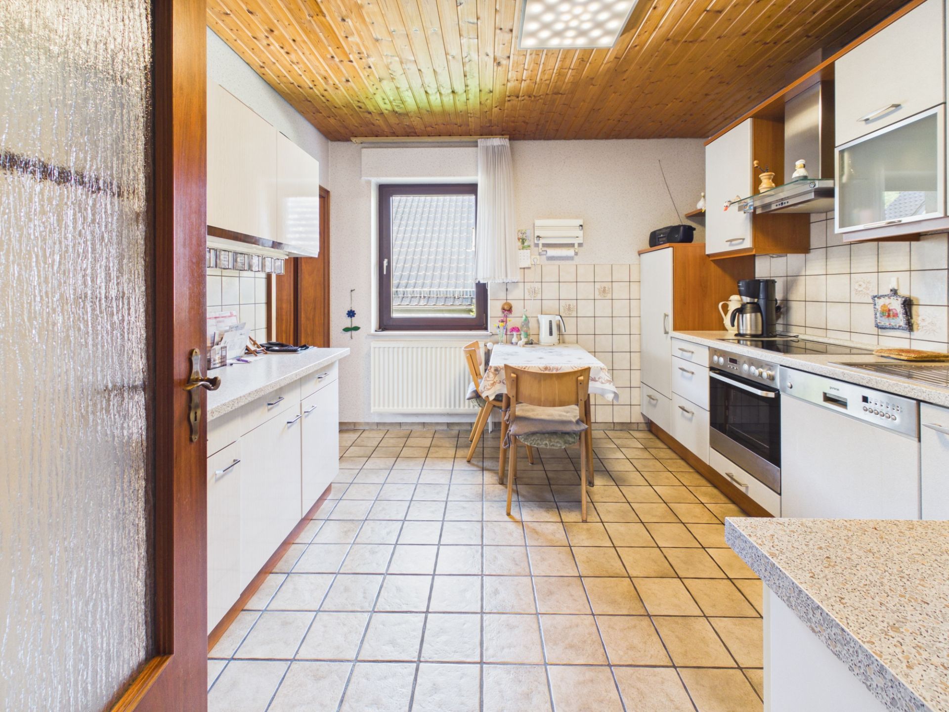 Helle Küche mit Esstisch, weißen Schränken, Fliesenboden und Holzdecke.