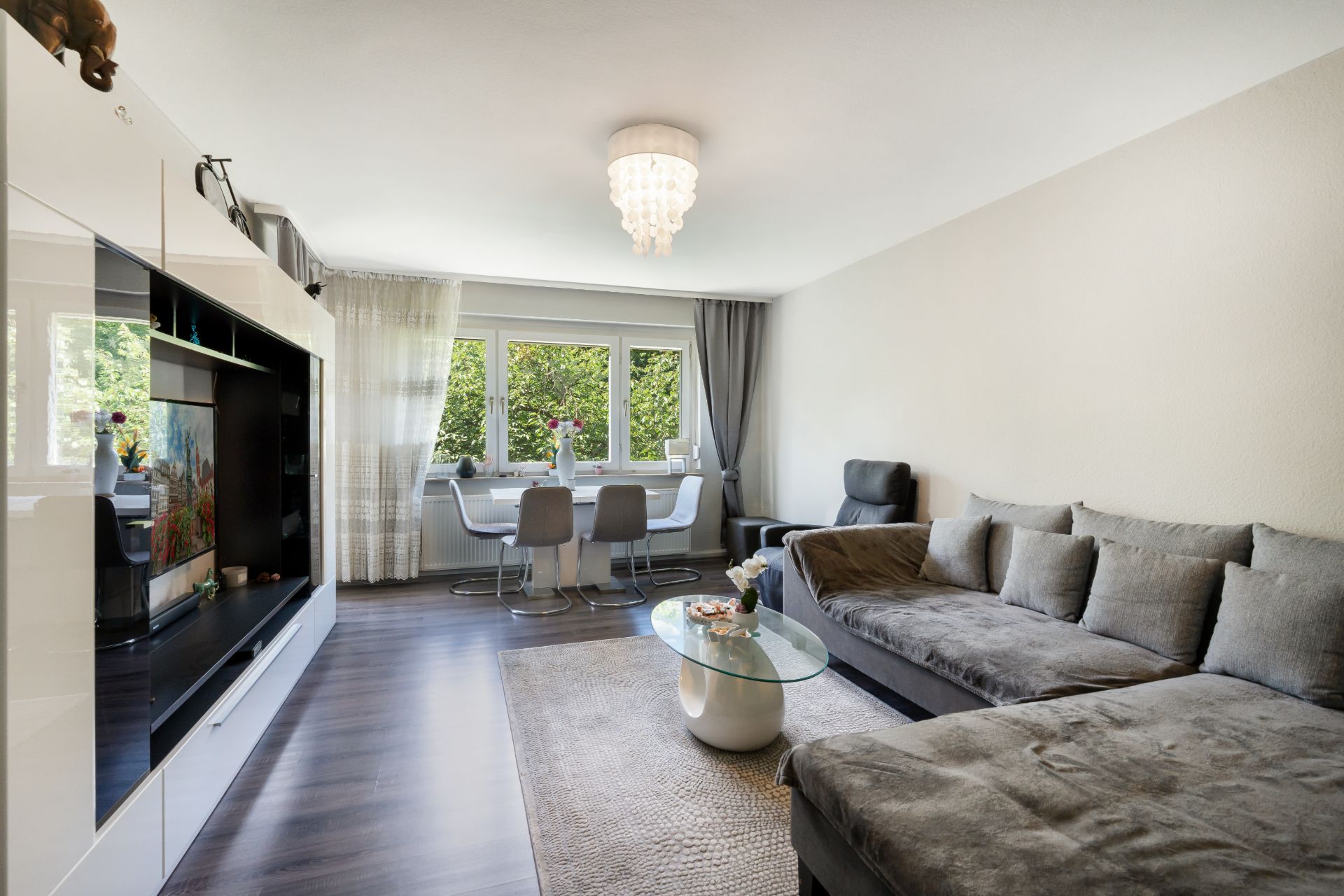 Modernes Wohnzimmer mit grauem Sofa, Esstisch, TV und großem Fenster mit Vorhängen.