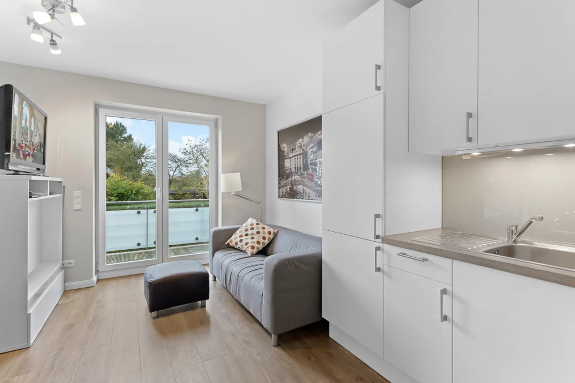 Wohnbereich mit grauem Sofa, Hocker, Küchenzeile und Zugang zu einem Balkon.