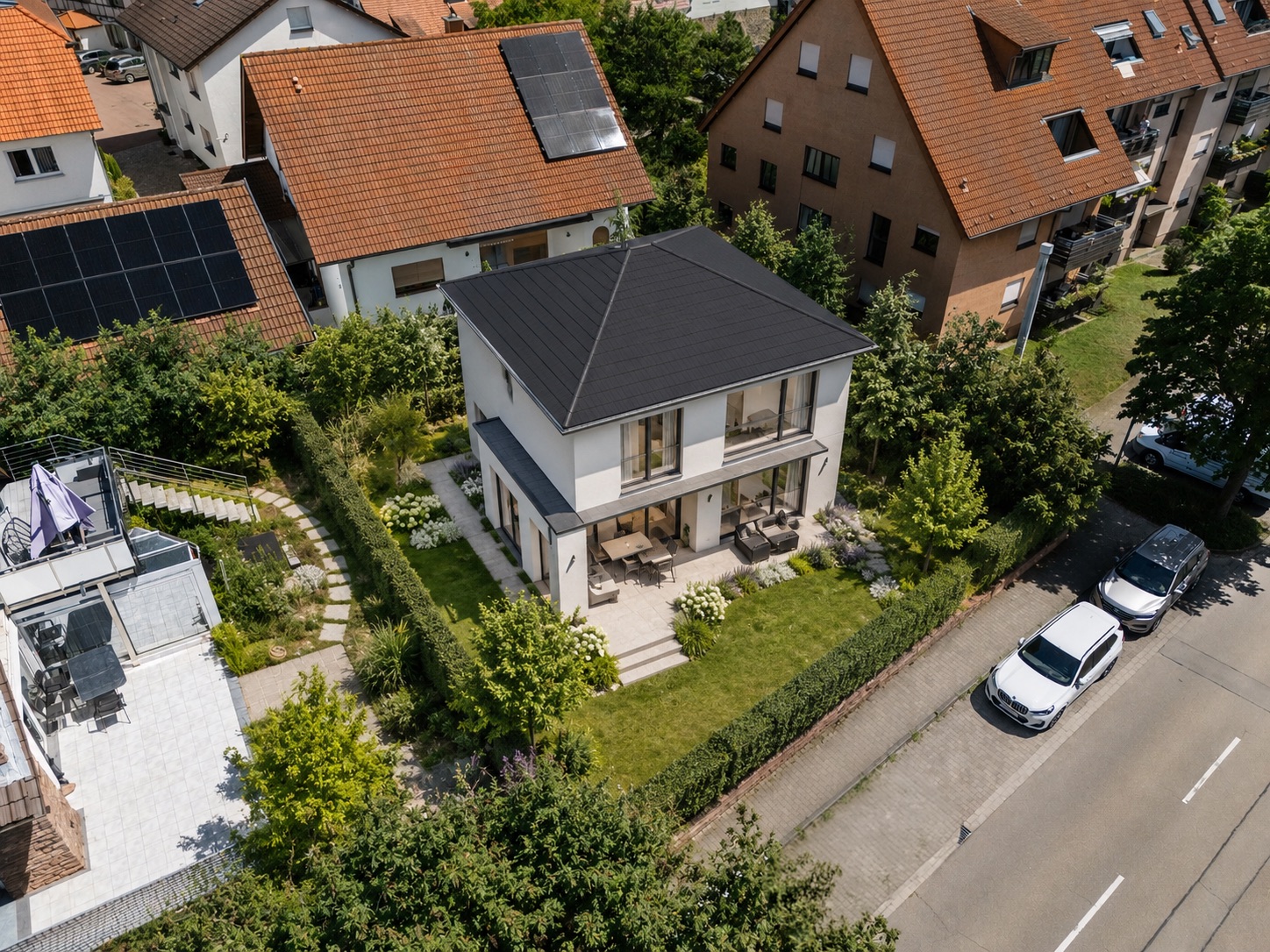 Modernes zweistöckiges Wohnhaus mit Garten, Terrasse und angrenzender Straße, umgeben von anderen Häusern.