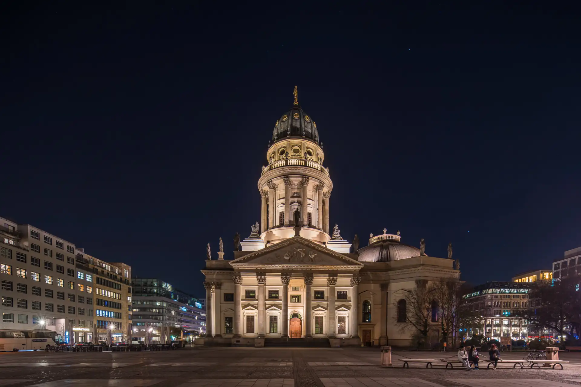 Beleuchtetes, historisches Gebäude mit Kuppel bei Nacht, umgeben von modernen Häusern.