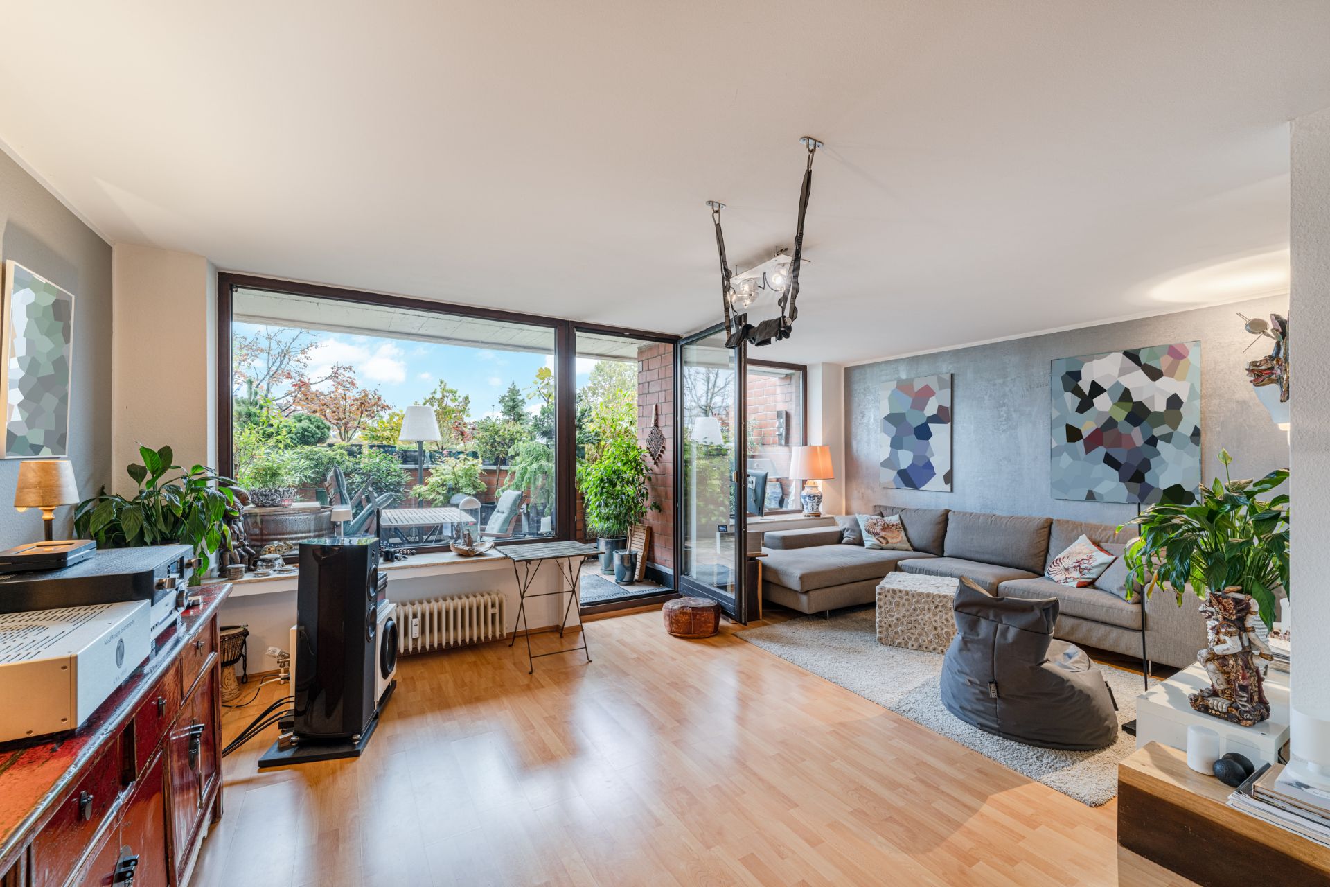 Helles Wohnzimmer mit Ecksofa, Pflanzen, Holzboden und Zugang zur Terrasse mit vielen Grünpflanzen.