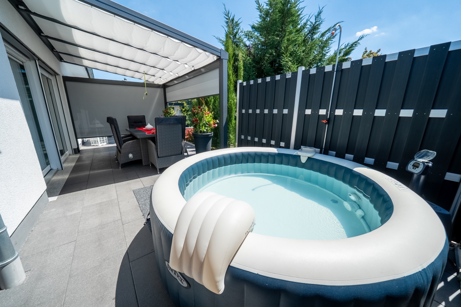 Auf einer Terrasse stehen ein aufblasbarer Whirlpool und ein Esstisch mit Stühlen unter einer Überdachung.
