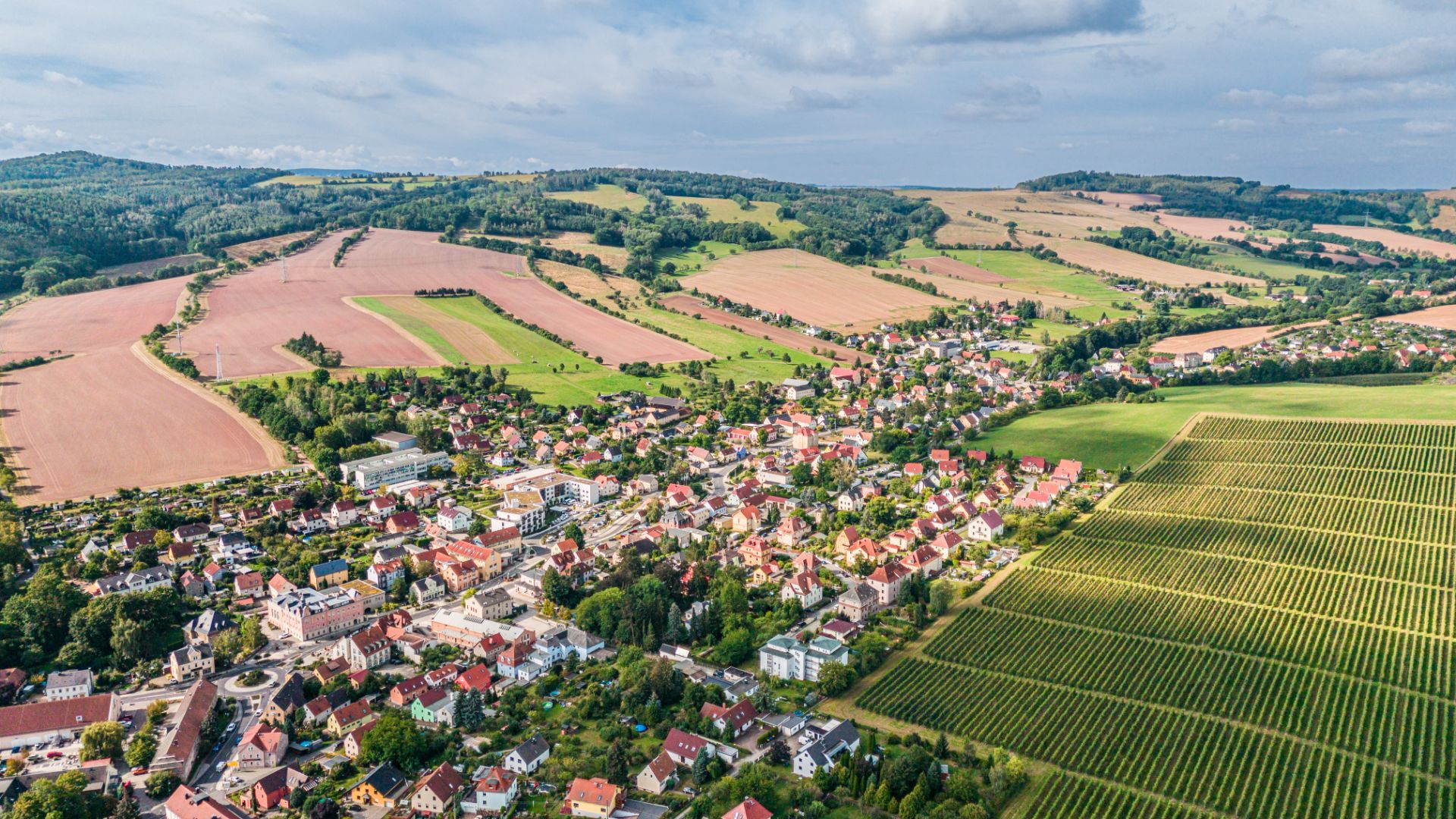 Luftaufnahme eines Dorfes mit Feldern und Wohnhäusern, umgeben von grünen Hügeln.
