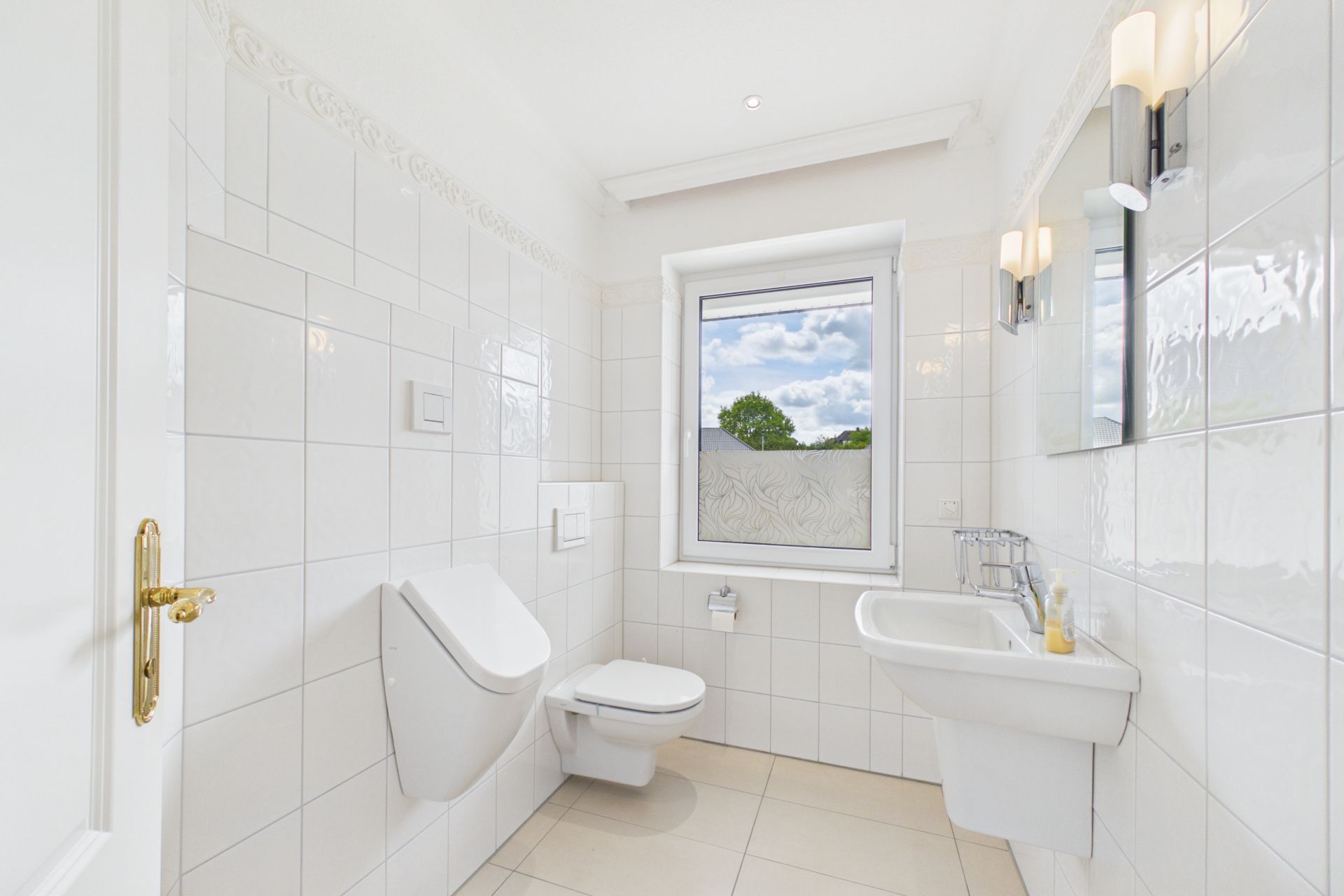 Helles Badezimmer mit Wandtoilette, Urinal, Waschbecken und Fenster.